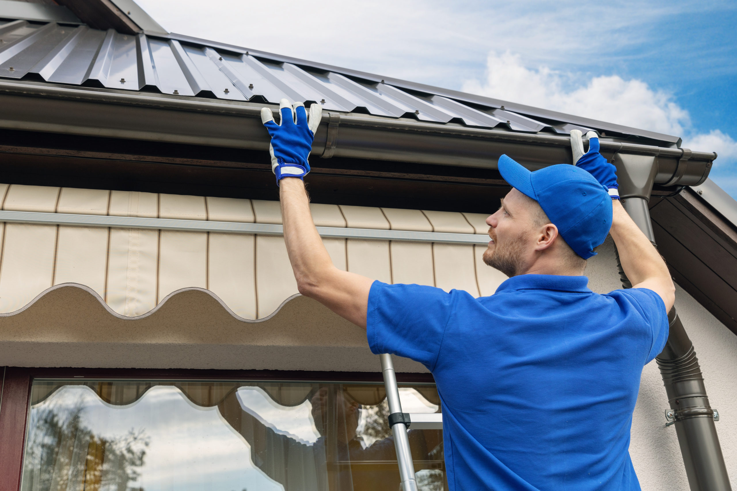 A man wearing a blue t-shirt is seen installing a house roof rain gutter. He is likely working on securing the gutter system to the edge of the roof, ensuring proper drainage of rainwater